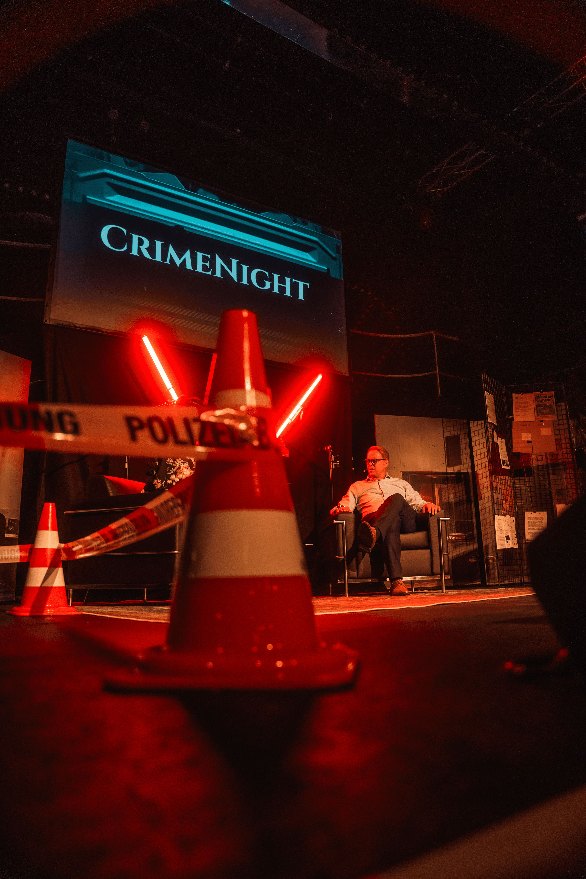 Ein Host sitzt im roten Licht auf der Bühne, im Vordergrund Leitkegel und Absperrband, dahinter die CrimeNight-Leinwand