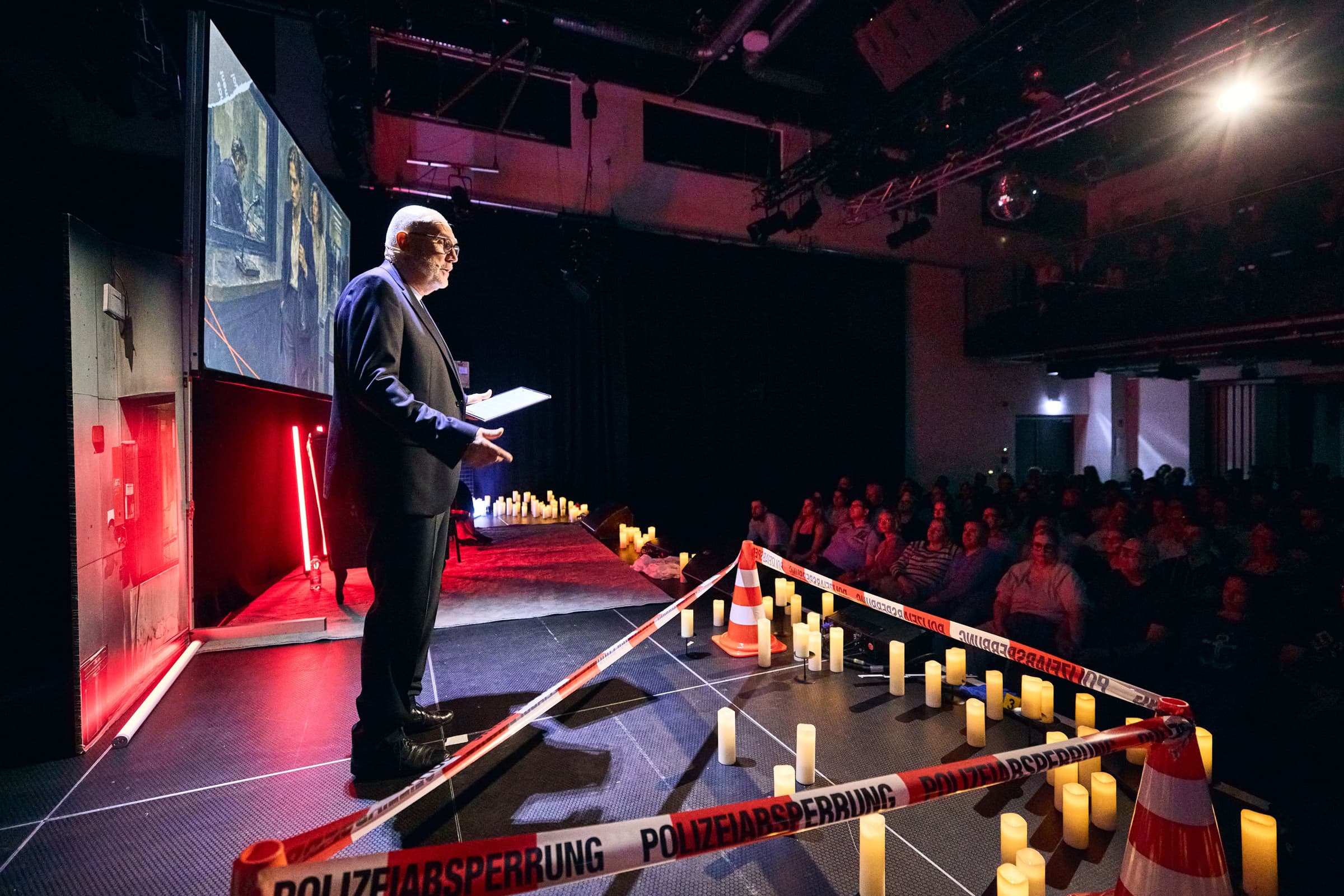 Ein Host der CrimeNight steht mit Notizen auf der abgedunkelten Bühne, davor Kerzen und rot-weißes Absperrband